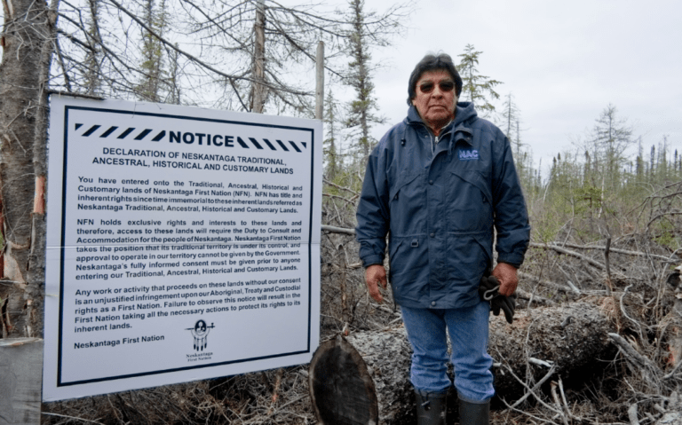 Former Chief of Neskantaga First Nation, Peter Moonias, stands next to a declaration of Neskantaga’s homelands, which community members have posted throughout their traditional territory to advise prospectors and mining companies. Photo by Allan Lissner/Neskantaga First Nation.